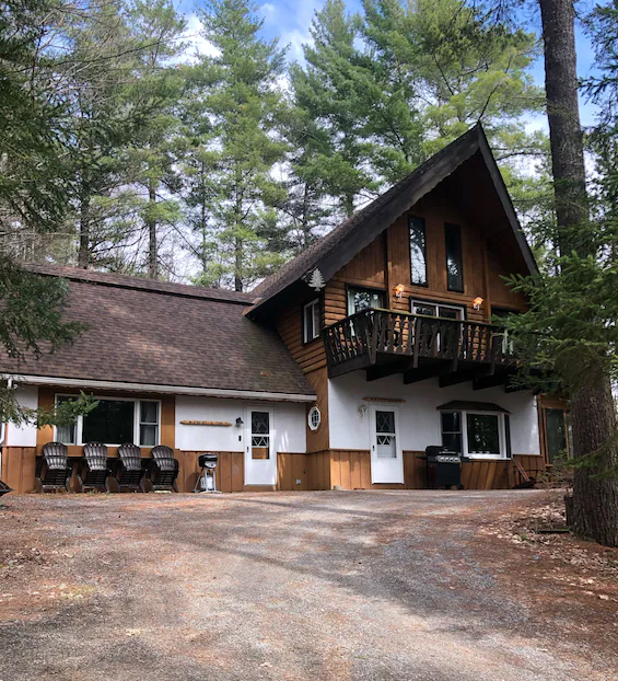 A picture of The Chalet at Jay, New York. At the end of a gravel road is a is a three story building with lots of wood accents, surrounded by trees.